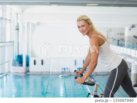 Woman in athletic wear pedaling stationary bike beside pool lanes in sports center, copy space Woman in athletic wear pedaling stationary bike beside pool lanes in sports center, copy space 135087890