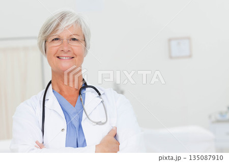 Senior female doctor standing smiling in exam room wearing lab coat stethoscope eyeglasses Senior female doctor standing smiling in exam room wearing lab coat stethoscope eyeglasses 135087910