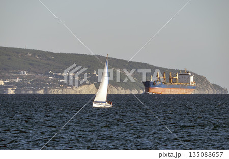 August 22 2025 Cargo ship in the background while a sailing yacht passes in the foreground near the port of Novorossiysk Russia. Maritime traffic on the Black Sea. 135088657