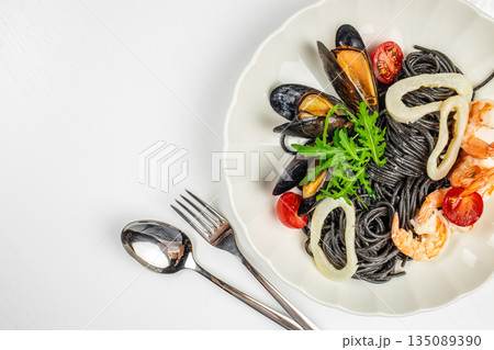 Black squid ink pasta with seafood, mussels, shrimp, calamari, and cherry tomatoes on a white plate. copy space, top view Black squid ink pasta with seafood, mussels, shrimp, calamari, and cherry tomatoes on a white plate. copy space, top view 135089390
