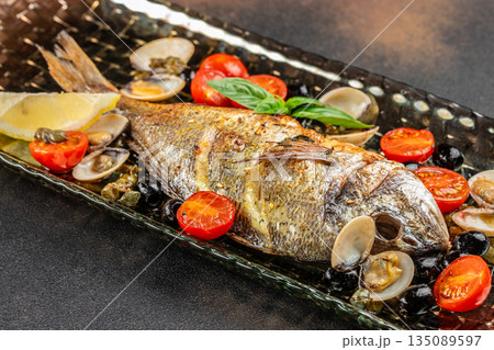 Baked whole fish with tomatoes olives and clams on a black plate. top view. copy space Baked whole fish with tomatoes olives and clams on a black plate. top view. copy space 135089597