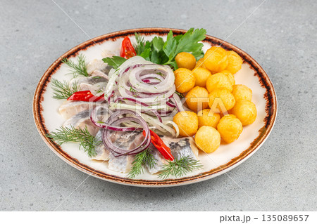 Pickled herring fillets served with crispy potato balls, red onion rings, fresh dill, parsley, and chili pepper on a ceramic plate 135089657