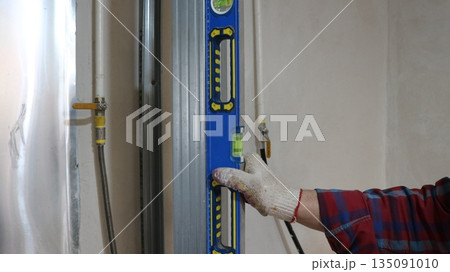 A worker in a checkered shirt and gloves holds a blue level on the surface of a vertically installed metal profile in a bright room with water and gas pipes, checking the structure in a technical room A worker in a checkered shirt and gloves holds a blue level on the surface of a vertically installed metal profile in a bright room with water and gas pipes, checking the structure in a technical room 135091010
