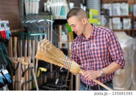 Male buyer carefully chooses broom in hardware store to clean yard of her house Male buyer carefully chooses broom in hardware store to clean yard of her house 135091393