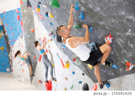 Atlete man grabbing ledges of artificial climbing wall in bouldering centre Atlete man grabbing ledges of artificial climbing wall in bouldering centre 135091420
