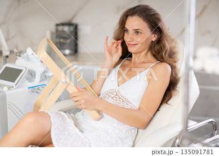 Positive woman examining her face after cosmetic procedure in medical room 135091781