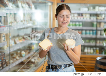Girl in bio food store holds cheese of different varieties in hands and is thinking about buying 135091940