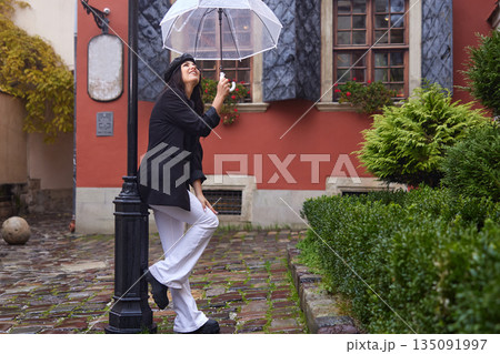 Fashionable woman, Woman outdoors, Rain clothing. In stylish black coat and beret, woman smiles while walking on rainy cobblestone street, holding transparent umbrella. Fashionable woman, Woman outdoors, Rain clothing. In stylish black coat and beret, woman smiles while walking on rainy cobblestone street, holding transparent umbrella. 135091997