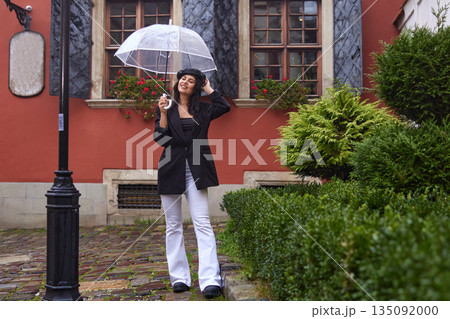Cobblestone street, Rain fashion, Umbrella rain. On rainy day, woman in stylish black coat and beret smiles while holding transparent umbrella and strolling along cobblestone street. Cobblestone street, Rain fashion, Umbrella rain. On rainy day, woman in stylish black coat and beret smiles while holding transparent umbrella and strolling along cobblestone street. 135092000