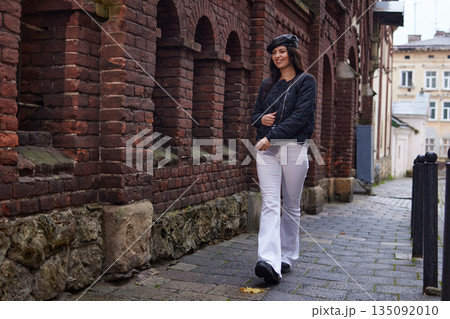 Casual smile, city stroll, fashion photography. Stylish woman walks past brick wall on rainy day, wearing black jacket and white pants and smiling casually. 135092010