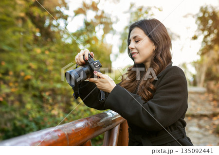 Young woman reviewing photos on her digital camera while standing in a sunny park. Warm autumn light, creative outdoor photography moment, lifestyle scene. 135092441