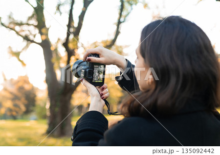 Young woman reviewing photos on her digital camera while standing in a sunny park. Warm autumn light, creative outdoor photography moment, lifestyle scene. 135092442