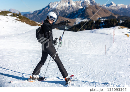Portrait young adult happy smiling woman skier wear black outfit overall stand snowy slope raised hand helmet goggles cold sunny winter day. Alpine resort mountain scenery blue sky Portrait young adult happy smiling woman skier wear black outfit overall stand snowy slope raised hand helmet goggles cold sunny winter day. Alpine resort mountain scenery blue sky 135093365