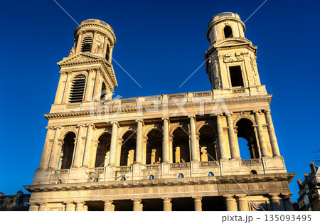 Church of Saint Sulpice stands in the 6th district of Paris, France. Historic stone facade features two mismatched towers and columns under a blue sky 135093495