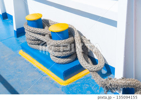 A detailed view of a thick rope tied around a blue bollard, located on a boat near a calm water body. Represents maritime activities, boating and nautical themes. High quality photo 135094155