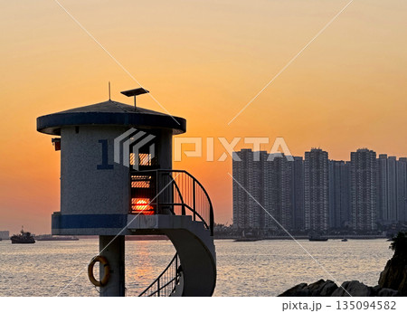Coastal observation tower with spiral staircase against a vibrant sunset and city skyline 135094582