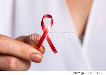 A doctor holds a red ribbon in their hand, symbolizing awareness for World Cancer Day on February 4th. The doctor wears a white coat and shows commitment to cancer awareness. 135094706
