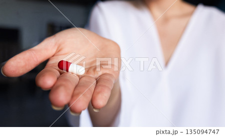 A young woman doctor in a white coat holds a red and white capsule in her right hand. The background is softly blurred, emphasizing the pill and the doctor. A young woman doctor in a white coat holds a red and white capsule in her right hand. The background is softly blurred, emphasizing the pill and the doctor. 135094747