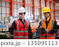 Two industrial workers in safety vests and helmets discussing operations while reviewing checklist on clipboard inside factory. Concept of team communication, planning and workplace safety. 135095113