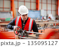 Industrial engineer wearing white safety helmet and red reflective vest operates heavy machinery in manufacturing plant, emphasizing focus, precision and hands on work in heavy factory. 135095127