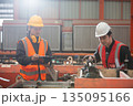Two factory workers in safety uniform inspecting machinery with clipboard in manufacturing plant. Industrial teamwork concept focusing on engineering process, precision and maintenance. 135095166