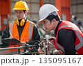 Two factory workers in safety uniform inspecting machinery with clipboard in manufacturing plant. Industrial teamwork concept focusing on engineering process, precision and maintenance. 135095167