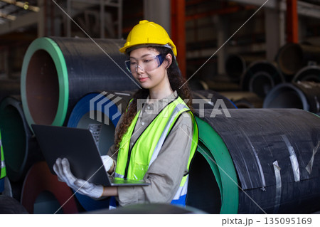 Female engineer inspects data on laptop inside metal sheet factory warehouse, focusing on production monitoring, inventory tracking and industrial workflow efficiency. 135095169