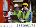 Two industrial engineers wearing safety helmets and reflective vests work together in factory using laptop, focusing on electrical system inspection, data analysis, and equipment monitoring. 135095172