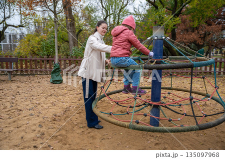 Mother Steadying Daughter On Spinning Roundabout Active Play With Supervision, Autumn Park, Jacket And Hat, Trusting Grip, Energetic Circle Of Motion Mother Steadying Daughter On Spinning Roundabout Active Play With Supervision, Autumn Park, Jacket And Hat, Trusting Grip, Energetic Circle Of Motion 135097968