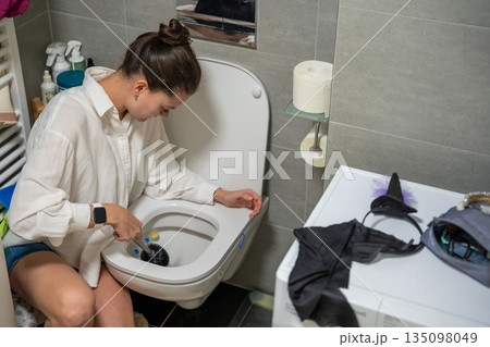 A woman cleans a toilet using a brush in a modern bathroom at home. Real life interior scene showing everyday household chores, hygiene routine and invisible domestic labor. 135098049