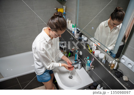 A woman washes her hands at a sink in a home bathroom. Real life interior scene showing everyday hygiene routine, personal care habit and domestic cleanliness. 135098097