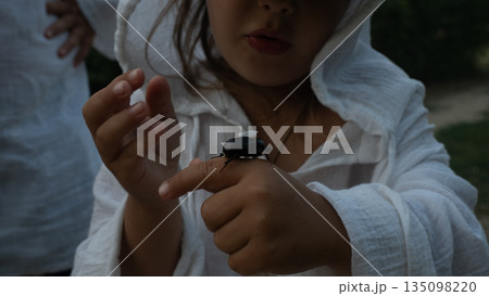 A child wearing a white hooded shirt carefully touches a large beetle crawling on his arm. The child displays an expression of surprise and curiosity about the insect in its natural outdoor setting. 135098220