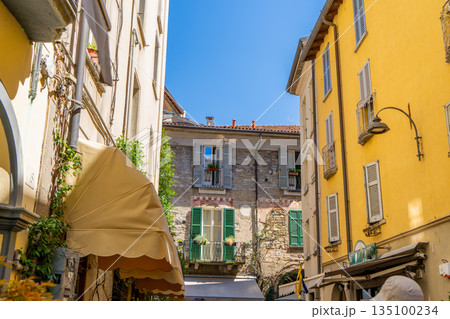 Multicolor windows balcony with shutters. Italy empty narrow street in summer day. Italian style facade architecture. Yellow wall Multicolor windows balcony with shutters. Italy empty narrow street in summer day. Italian style facade architecture. Yellow wall 135100234