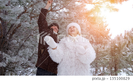 Joyful Couple Plays In Winter Snow, Rejoicing Pair Under Frosted Pine Trees During Snowstorm, Enthusiastic Duo Raises Arms Amidst Snowfall Beneath Icy Pine Branches On Winter Afternoon 135100829