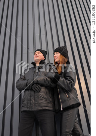 Fashionforward Pair In Dark Clothing Poses Near Architectural Background, Two Individuals Donning Black Jackets Stand Confidently Against Contemporary Striped Wall Showcasing Urban Style 135100862