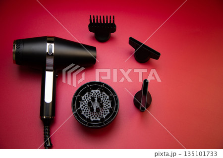 Black hair dryer, next to it there are various drying attachments - a comb, a diffuser and classic nozzles of different diameters, on a pink background, top view. High quality photo Black hair dryer, next to it there are various drying attachments - a comb, a diffuser and classic nozzles of different diameters, on a pink background, top view. High quality photo 135101733