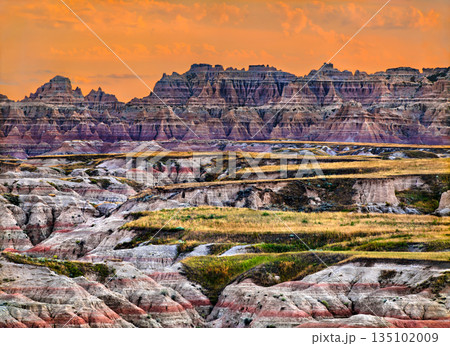 Big Badlands Overlook in Badlands National Park, South Dakota, USA. Scenic view features colorful eroded rock formations and grassy layers under a bright orange sunset sky 135102009