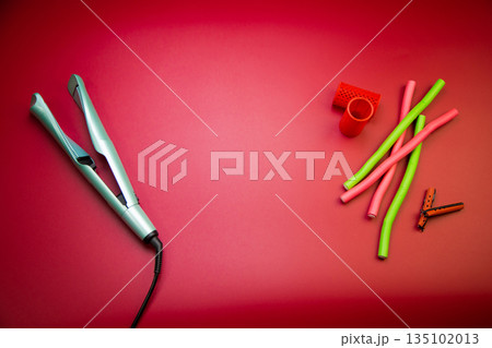 Top view of a green hair straightener with a swirling ceramic canvas, next to which there are curlers of different shapes and sizes on a pink background with space for text. The concept of styling Top view of a green hair straightener with a swirling ceramic canvas, next to which there are curlers of different shapes and sizes on a pink background with space for text. The concept of styling 135102013