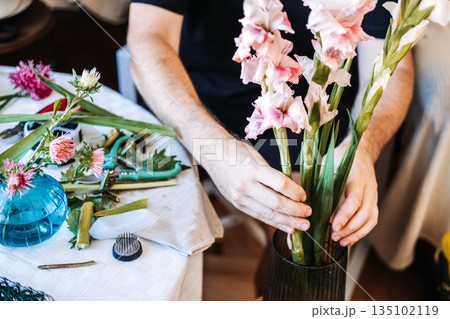 Hands hold large pink and white gladiolus stems above work table covered in botanical trimmings and floral tools. DIY floral design, creative skill-building, hobbyist floriculture. Hands hold large pink and white gladiolus stems above work table covered in botanical trimmings and floral tools. DIY floral design, creative skill-building, hobbyist floriculture. 135102119