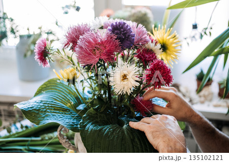 Hands hold finished floral arrangement in stone urn featuring colorful asters and large green hosta leaves. Floral design class, DIY flower arranging, floristry workshop. 135102121
