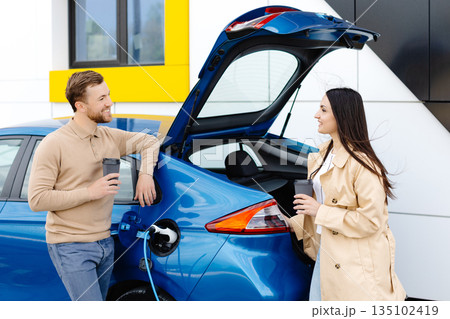Young couple man and woman traveling by electric car having stop at charging station. Boyfriend plugging in cable to charge. 135102419
