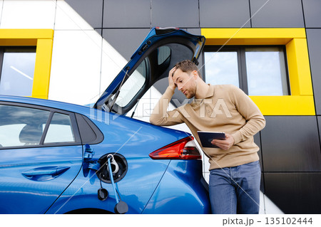 A young man is charging an electric car while standing at a station and holding a tablet 135102444