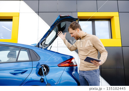 A young man is charging an electric car while standing at a station and holding a tablet A young man is charging an electric car while standing at a station and holding a tablet 135102445