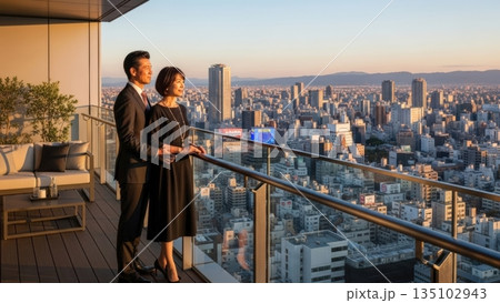 夕景を背にタワーマンションのベランダから将来を眺める成功者のカップル 135102943