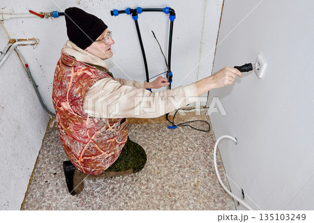 Man inserts electrical plug of heating cable for water pipe into socket in cold utility room with incoming water pipe. Man inserts electrical plug of heating cable for water pipe into socket in cold utility room with incoming water pipe. 135103249