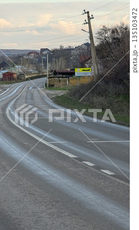 Wet Country Road Heading Into Village With Weathered Markings And Surrounding Natural Features Wet Country Road Heading Into Village With Weathered Markings And Surrounding Natural Features 135103472