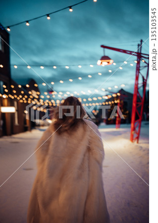 Woman walks on snowy street with string lights during evening hours in a festive setting 135103545