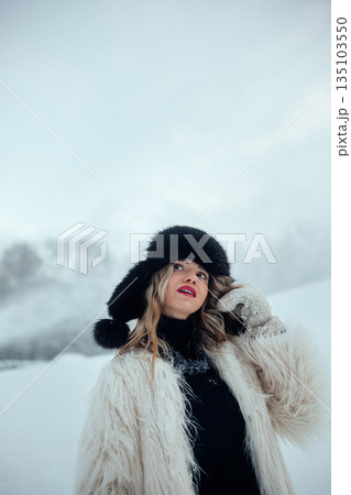 Woman poses in winter attire in snowy landscape with soft clouds in the sky 135103550