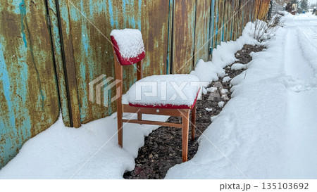 Old red upholstered wooden chair heavily covered in fresh snow, abandoned outdoors next to a rusty wall and a blue fence during a cold winter day. Old red upholstered wooden chair heavily covered in fresh snow, abandoned outdoors next to a rusty wall and a blue fence during a cold winter day. 135103692