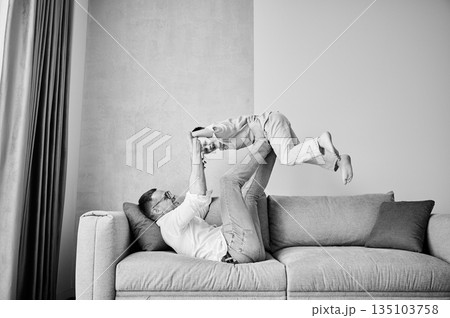 Joyful father lifting happy little son at home. Carefree father and son weekend. Happy young father lying on couch and fooling around with kid. Black and white image. Joyful father lifting happy little son at home. Carefree father and son weekend. Happy young father lying on couch and fooling around with kid. Black and white image. 135103758